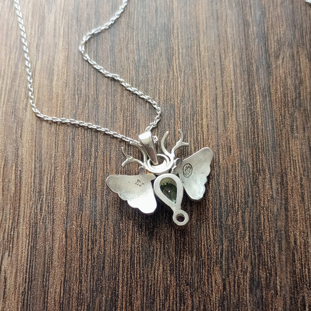 The back of a small moth pendant. The wings are formed by two months surrounding a central pear shaped green sapphire below which there is a small pink sapphire. Antlers replace antennas. Stamped BJ for the designer Brydie Jayne and 925 for Sterling Silver. Displayed on a wood like surface.