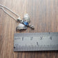 A small moth pendant. The wings are formed by two months surrounding a central claw set oval cabochon sapphire below which there is a small free moving pear shaped rainbow Moonstone. A Cresent moon represents antennas in the middle. Displayed on a wood like surface next to a ruler for scale.