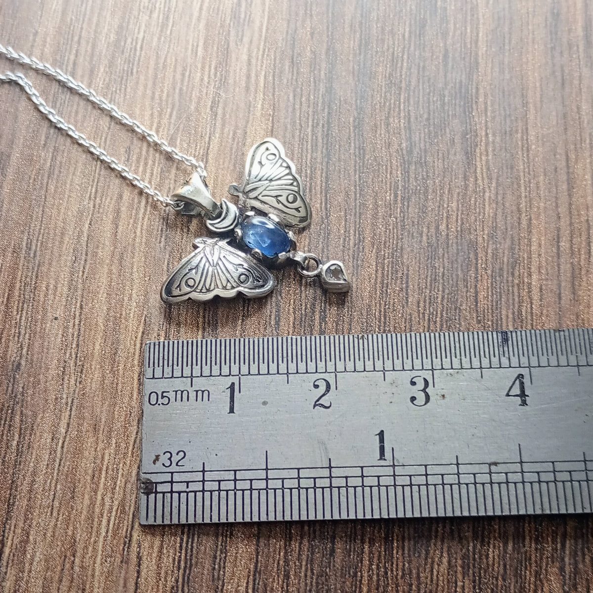 A small moth pendant. The wings are formed by two months surrounding a central claw set oval cabochon sapphire below which there is a small free moving pear shaped rainbow Moonstone. A Cresent moon represents antennas in the middle. Displayed on a wood like surface next to a ruler for scale.