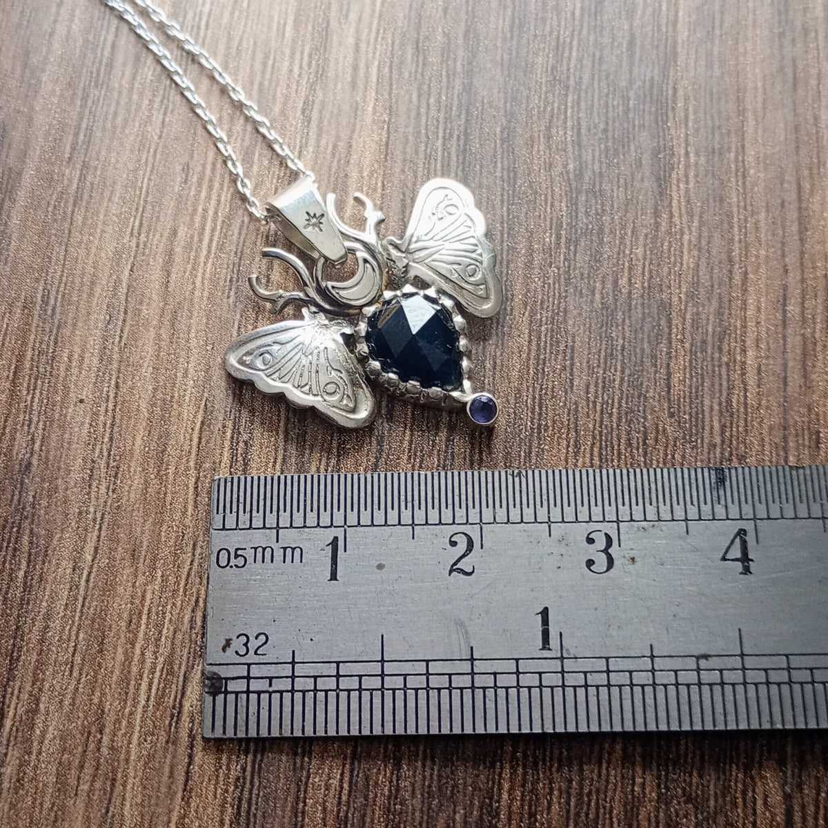 Silver Moth composed of two moth around an inverted pear shaped dark blue sapphire with antlers in places of antennas. Displayed on a wood look background with ruler for scale.