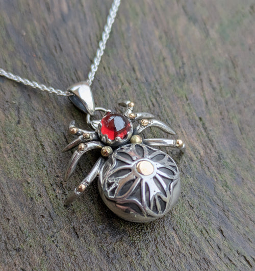 A detailed gothic rose window inspired spider made from Sterling Silver and 9ct yellow Gold featuring a blood-red garnet cabochon as the head of the spider. Each leg has a gold ball on where the leg bends and the spiders abdomen has a meticulously hand crafted gothic rose design.