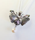A silver moth pendant made up of two months meeting at the top surrounding a claw set amethyst cabochon with a rope twist around it. The moth has antlers like a deer that include a lead motif and two small peridot stones. A pearl dangles from the tail of the moth and the whole piece is displayed on a silver chain with a white background.
