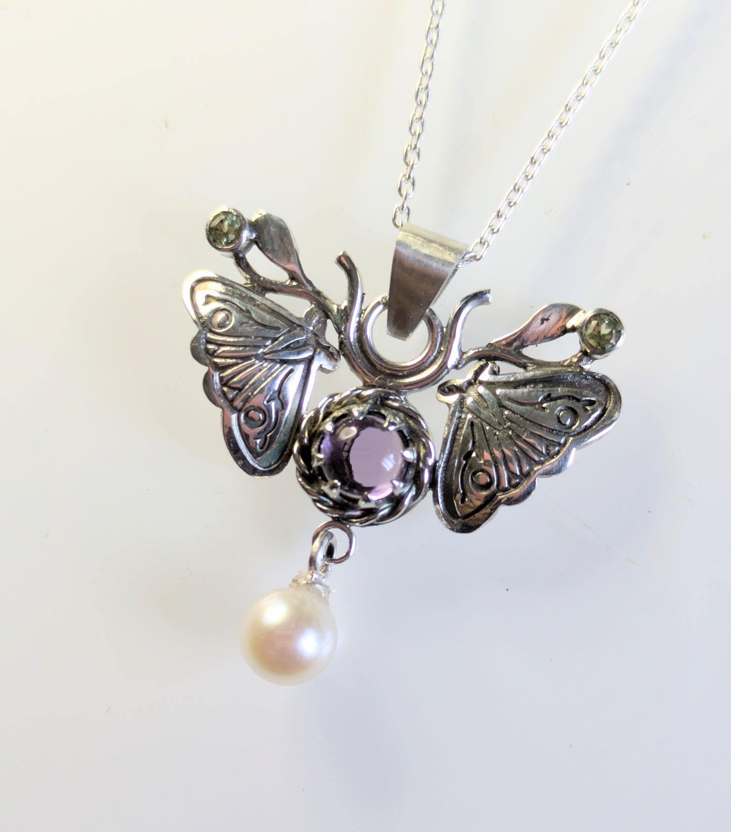 A silver moth pendant made up of two months meeting at the top surrounding a claw set amethyst cabochon with a rope twist around it. The moth has antlers like a deer that include a lead motif and two small peridot stones. A pearl dangles from the tail of the moth and the whole piece is displayed on a silver chain with a white background.