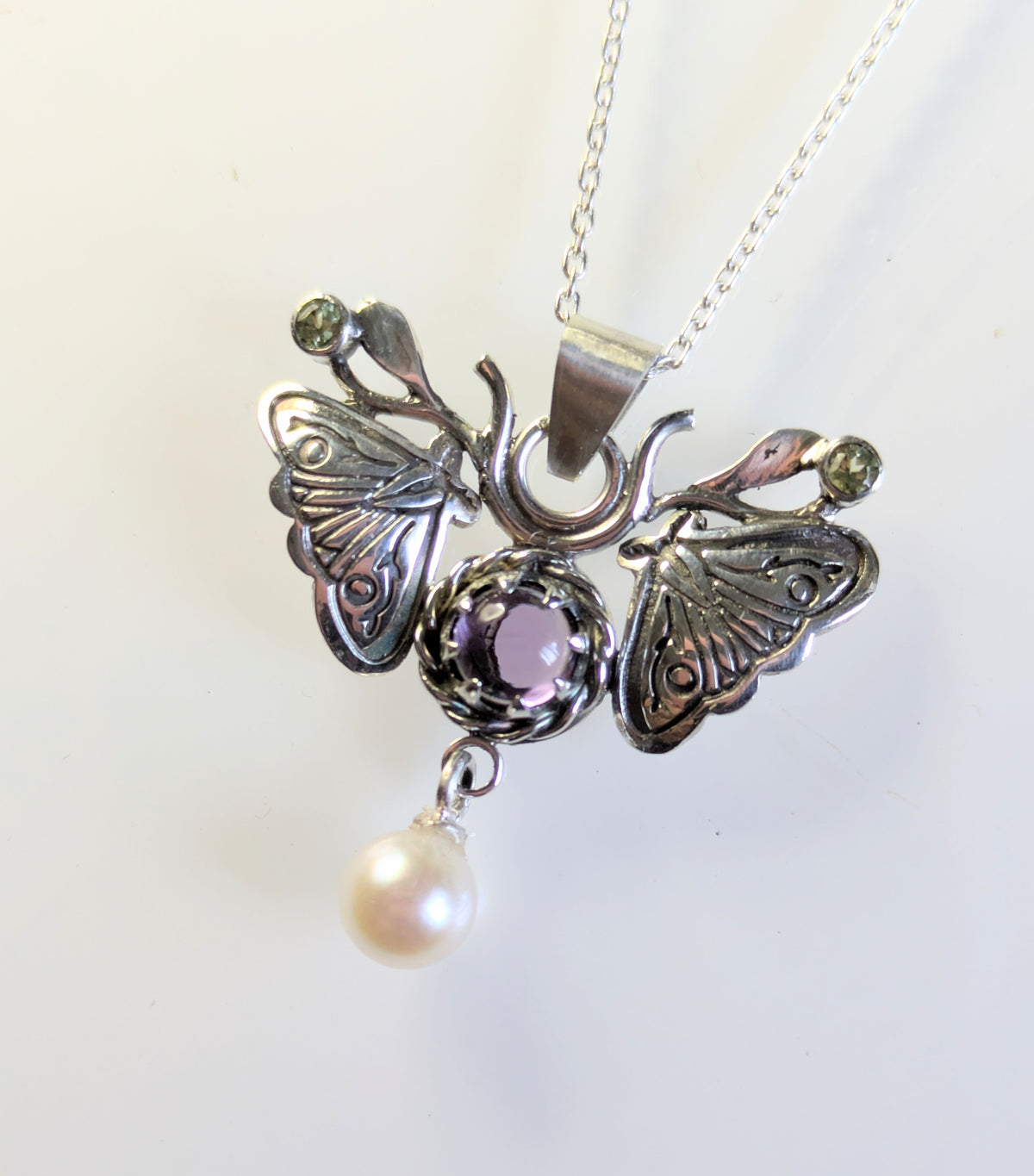 A silver moth pendant made up of two months meeting at the top surrounding a claw set amethyst cabochon with a rope twist around it. The moth has antlers like a deer that include a lead motif and two small peridot stones. A pearl dangles from the tail of the moth and the whole piece is displayed on a silver chain with a white background.