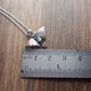 A small moth pendant. The wings are formed by two months surrounding a central claw set pake blue oval sapphire below which there is a small free moving round stone. A Cresent moon represents antennas in the middle. Displayed on a wood like surface next to a ruler for scale.