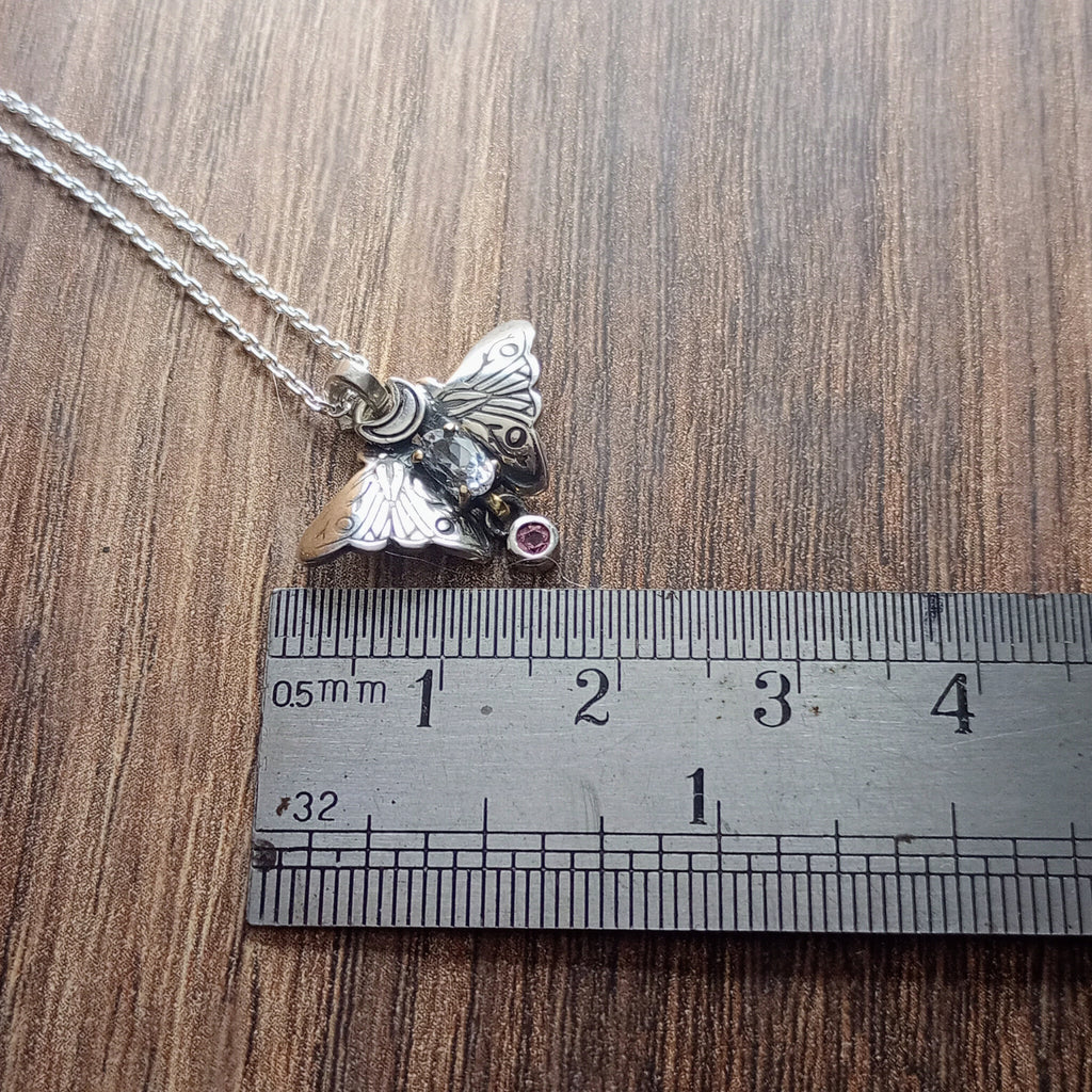 A small moth pendant. The wings are formed by two months surrounding a central claw set pake blue oval sapphire below which there is a small free moving round stone. A Cresent moon represents antennas in the middle. Displayed on a wood like surface next to a ruler for scale.