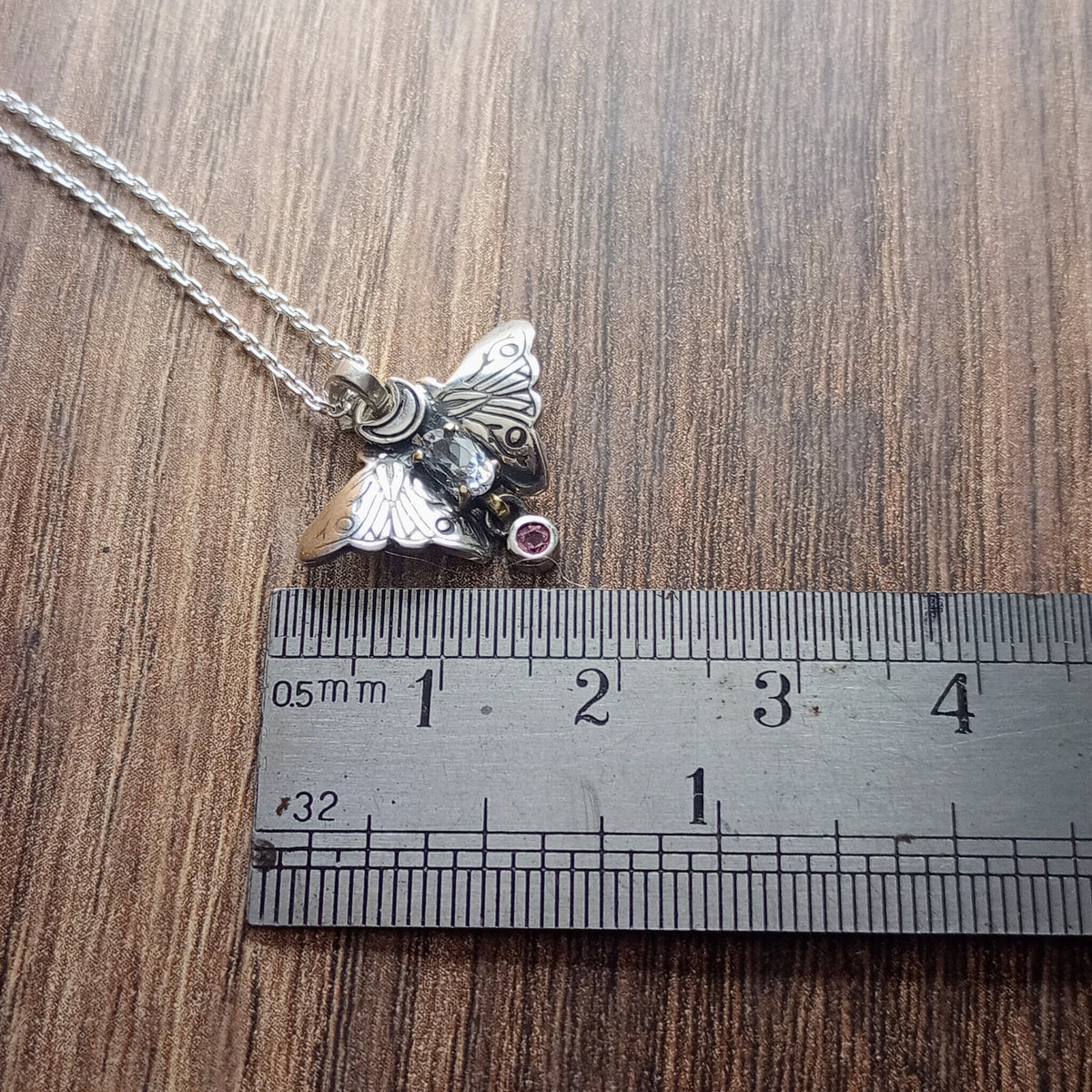 A small moth pendant. The wings are formed by two months surrounding a central claw set pake blue oval sapphire below which there is a small free moving round stone. A Cresent moon represents antennas in the middle. Displayed on a wood like surface next to a ruler for scale.
