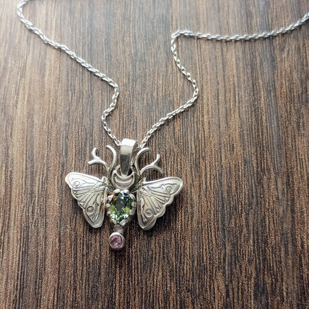 A small moth pendant. The wings are formed by two months surrounding a central claw set pear shaped green sapphire below which there is a small pink sapphire. Antlers replace antennas with a Cresent moon in the middle. Displayed on a wood like surface.