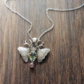 A small moth pendant. The wings are formed by two months surrounding a central claw set pear shaped green sapphire below which there is a small pink sapphire. Antlers replace antennas with a Cresent moon in the middle. Displayed on a wood like surface.