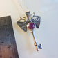 Silver key-shaped pendant with moth design and purple gemstone on a white background, next to a ruler for scale.