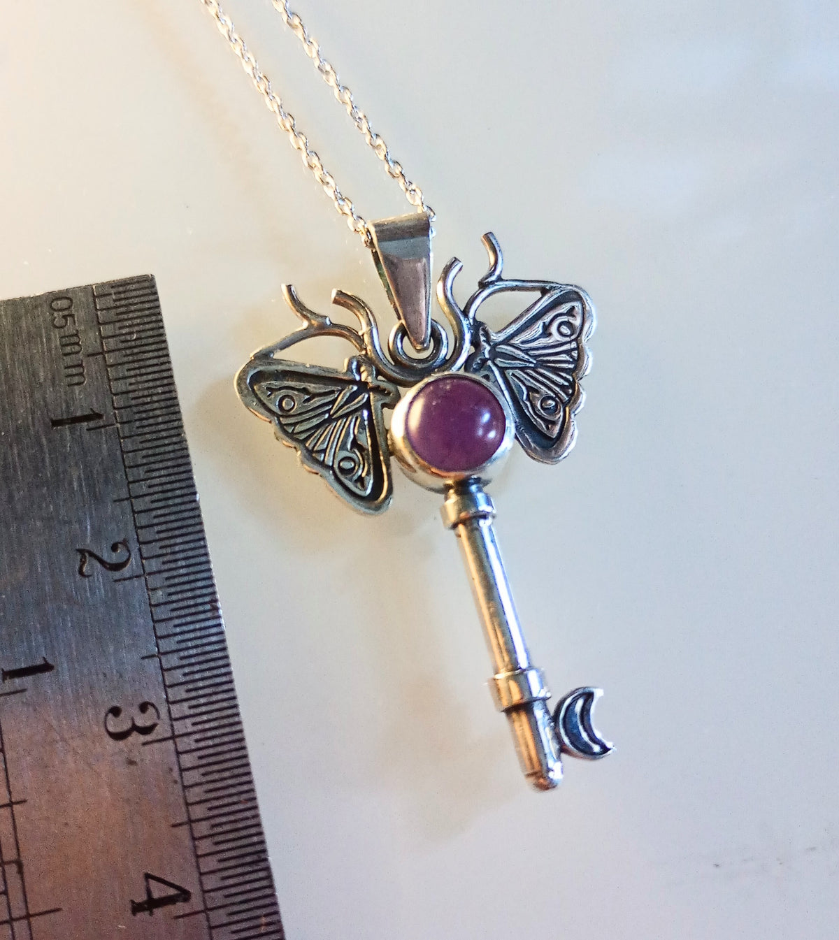 Silver key-shaped pendant with moth design and purple gemstone on a white background, next to a ruler for scale.