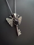 Silver and onyx moth key pendant necklace on a dark background. The piece has moon and a star on the key parts and deer antlers as antennas .