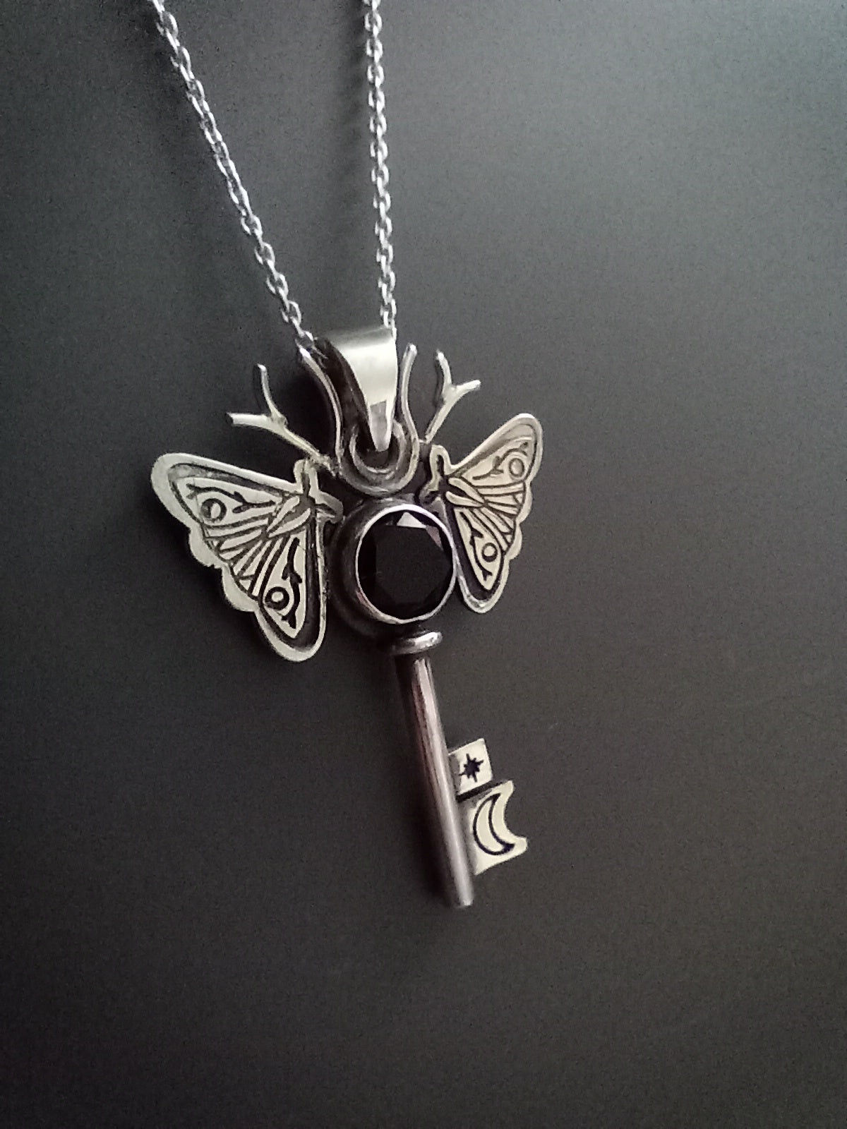 Silver and onyx moth key pendant necklace on a dark background. The piece has moon and a star on the key parts and deer antlers as antennas .