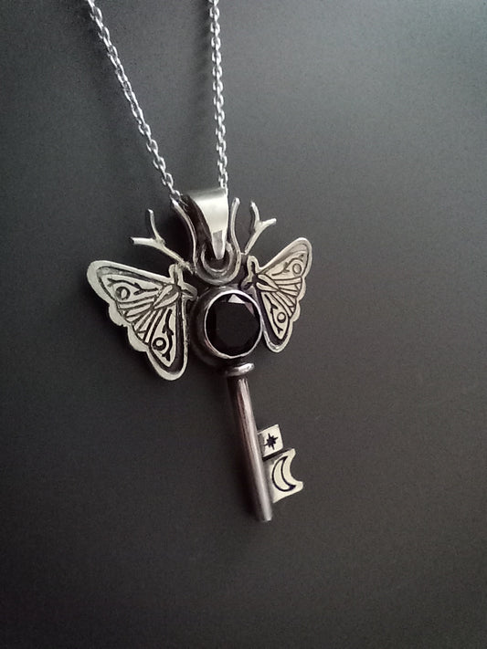 Silver and onyx moth key pendant necklace on a dark background. The piece has moon and a star on the key parts and deer antlers as antennas .