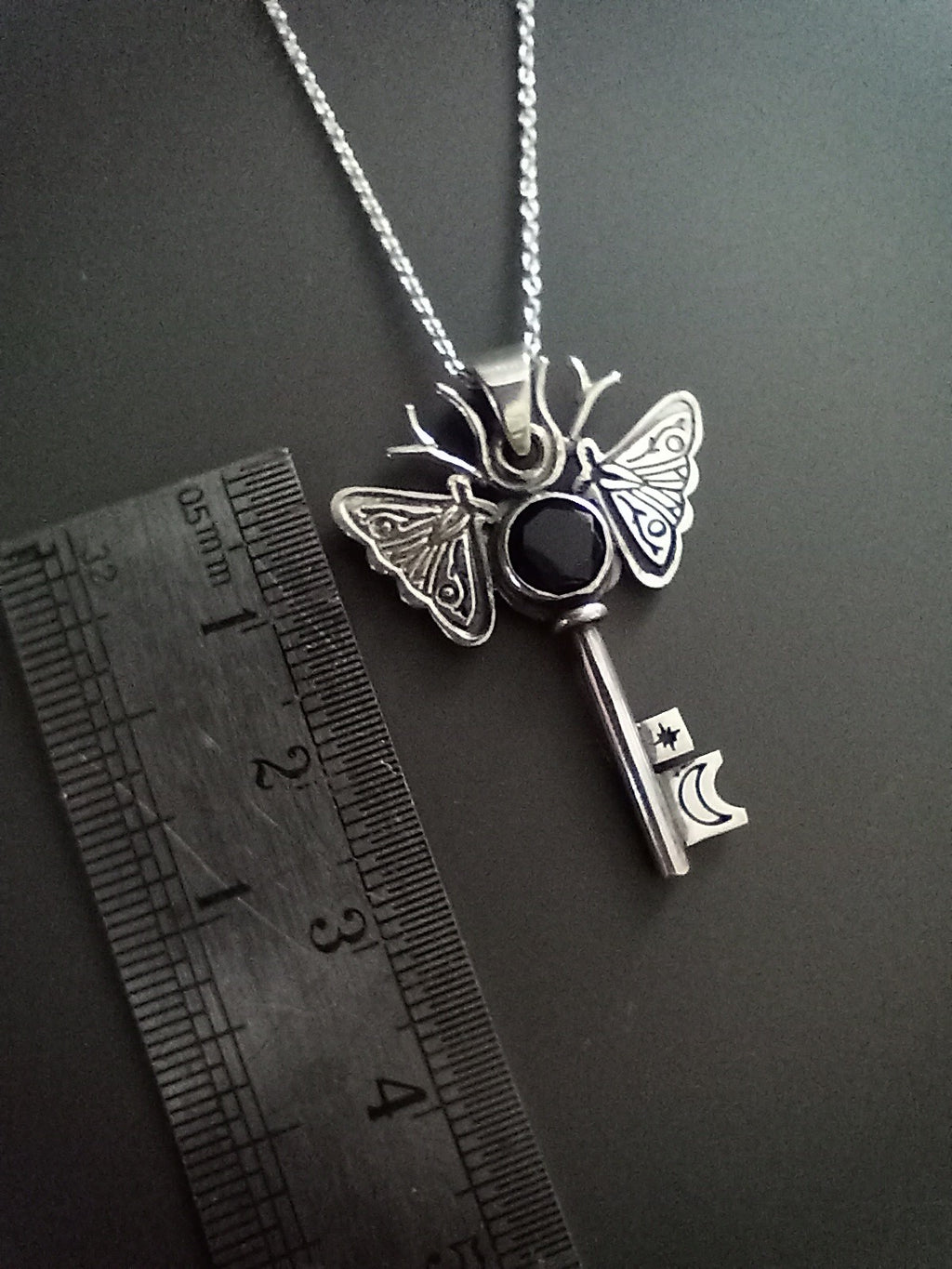 Silver and onyx moth key pendant necklace on a dark background. The piece has moon and a star on the key parts and deer antlers as antennas . Ruler for scale.