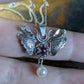 A silver moth pendant made up of two months meeting at the top surrounding a claw set amethyst cabochon with a rope twist around it. The moth has antlers like a deer that include a lead motif and two small peridot stones. A pearl dangles from the tail of the moth and the whole piece is displayed on a silver chain and is resting over fingers.