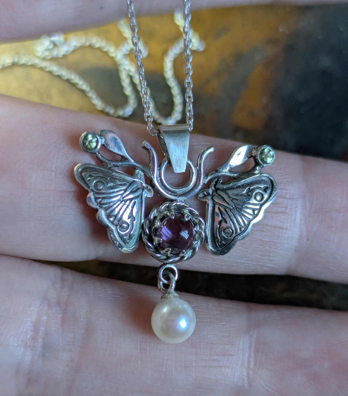 A silver moth pendant made up of two months meeting at the top surrounding a claw set amethyst cabochon with a rope twist around it. The moth has antlers like a deer that include a lead motif and two small peridot stones. A pearl dangles from the tail of the moth and the whole piece is displayed on a silver chain and is resting over fingers.