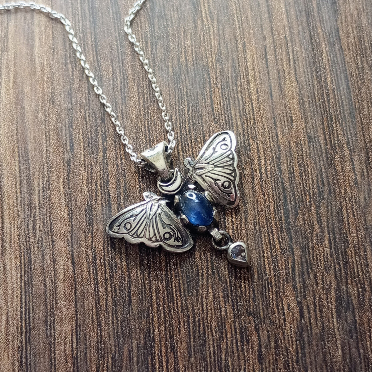 A small moth pendant. The wings are formed by two months surrounding a central claw set oval cabochon sapphire below which there is a small free moving pear shaped rainbow Moonstone. A Cresent moon represents antennas in the middle. Displayed on a wood like surface.
