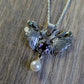 A silver moth pendant made up of two months meeting at the top surrounding a claw set amethyst cabochon with a rope twist around it. The moth has antlers like a deer that include a lead motif and two small peridot stones. A pearl dangles from the tail of the moth and the whole piece is displayed on a silver chain with a wood look background 