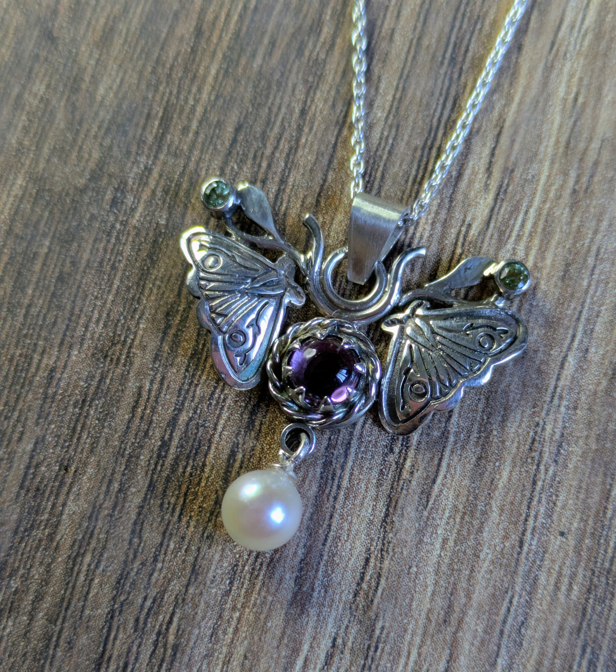 A silver moth pendant made up of two months meeting at the top surrounding a claw set amethyst cabochon with a rope twist around it. The moth has antlers like a deer that include a lead motif and two small peridot stones. A pearl dangles from the tail of the moth and the whole piece is displayed on a silver chain with a wood look background 