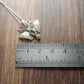 A small moth pendant. The wings are formed by two months surrounding a central claw set pear shaped green sapphire below which there is a small pink sapphire. Antlers replace antennas with a Cresent moon in the middle. Displayed on a wood like surface next to a ruler for scale.