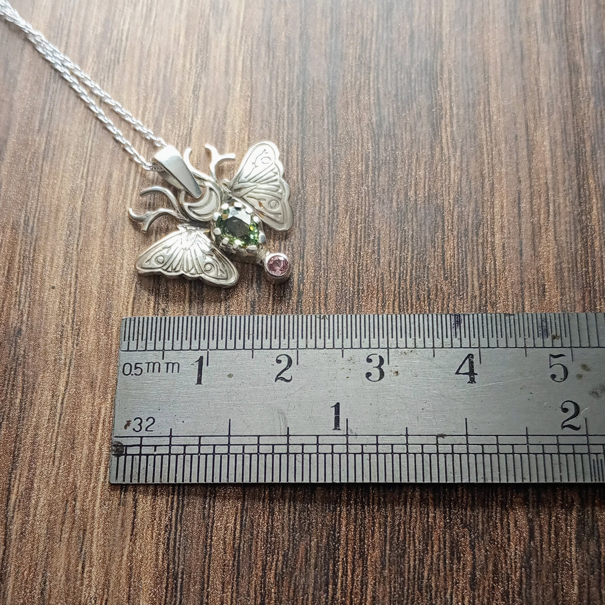A small moth pendant. The wings are formed by two months surrounding a central claw set pear shaped green sapphire below which there is a small pink sapphire. Antlers replace antennas with a Cresent moon in the middle. Displayed on a wood like surface next to a ruler for scale.