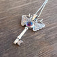Silver key-shaped pendant with moth wings and purple gemstone on a wooden surface
