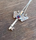 Silver key-shaped pendant with moth wings and purple gemstone on a wooden surface