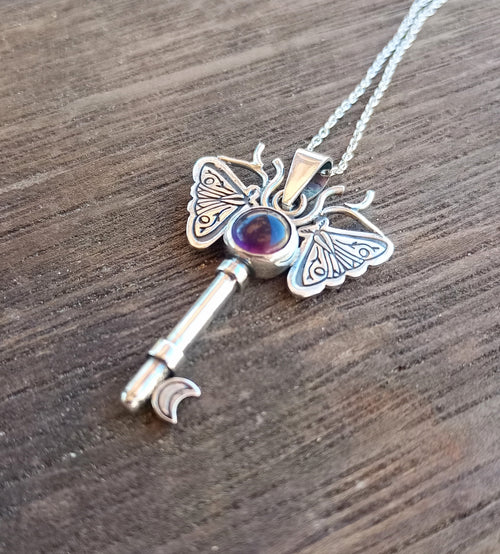 Silver key-shaped pendant with moth wings and purple gemstone on a wooden surface