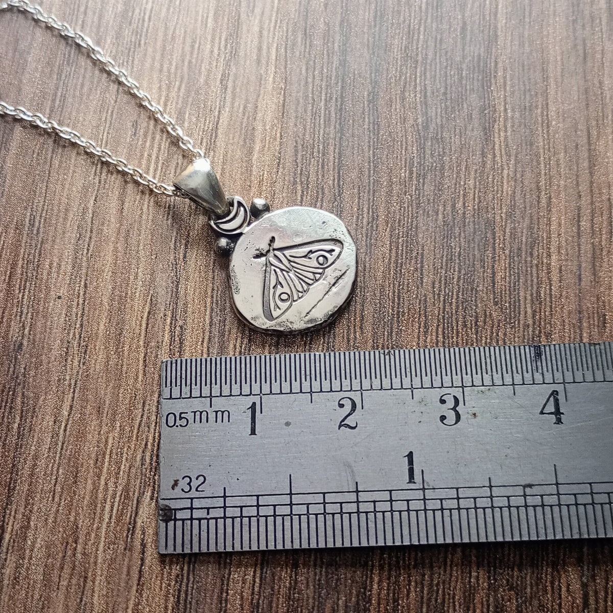 Oval shaped silver pendant with aged appearance stamped with moth design. Accented with a moon at top. Displayed on wood look background with ruler for scale.